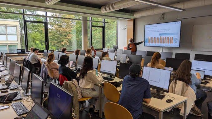 A group of researchers sitting at computer workstations in a bright seminar room, listening to a presentation on FReDA’s survey design shown on a large screen at the front of the room.