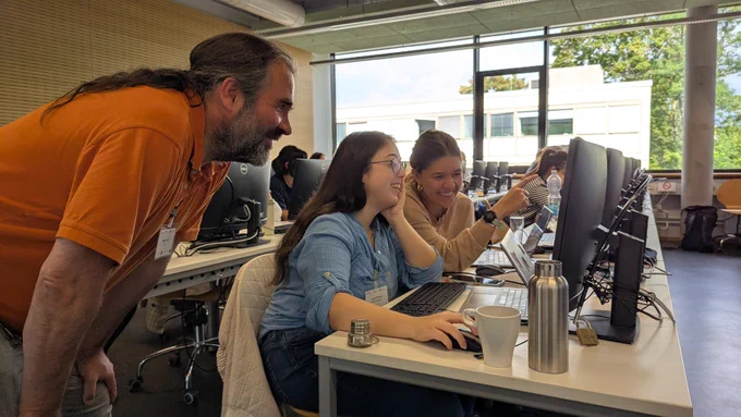 Two young researchers smiling while working together at a computer, supported by an instructor leaning in to help.