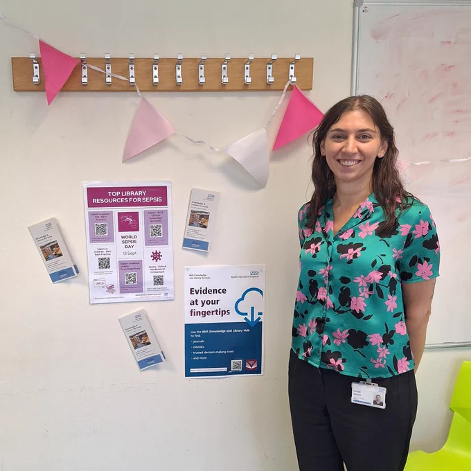 Librarian stood in front of display of library posters and pink banner for World Sepsis Day