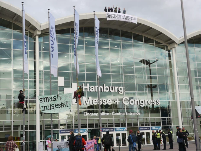 Zu sehen ist der Eingang zu den Messehallen in Hamburg. Auf zwei Fahnenmasten auf dem Vorplatz sind zwei Menschen hochgeklettert und haben ein Transpi gespannt auf dem "Smash Cruiseshit" steht. Auf dem Dach der Messehallen Stehen Menschen und haben ein Banner befestigt auf dem "Kreuzfahrt Stoppen!" drauf steht.