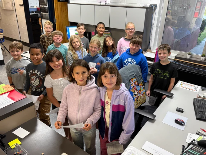 15 students stand in the front office and smile at the camera before they sing their song. they are huddled close together. 
