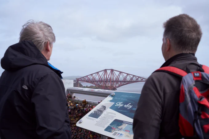 Two walkers looking towards the Forth Bridge from the tourist sign on the Forth Road Bridge.  