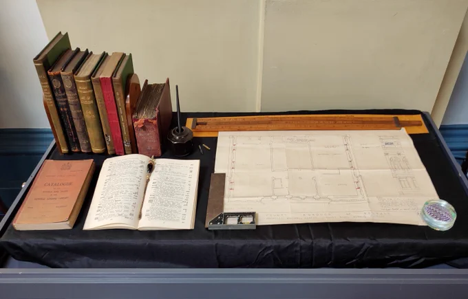 A collection of vintage books, an open catalogue, ink pot with a quill, and architectural blueprints displayed on a black cloth-covered table.