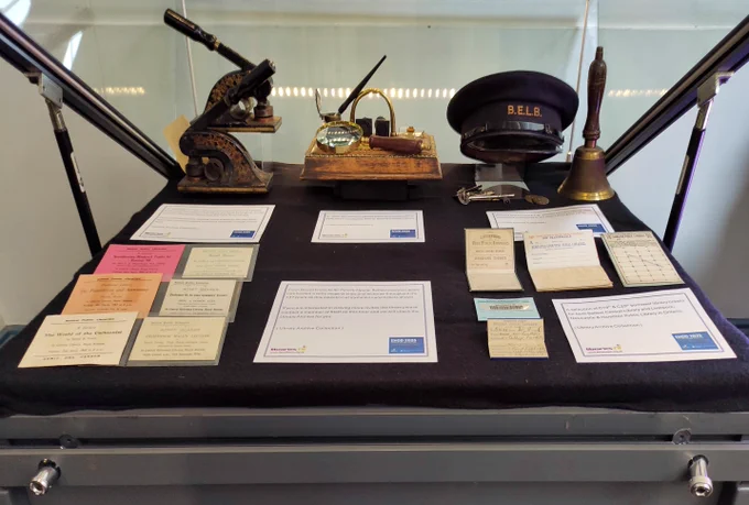Display case featuring vintage library tools including a magnifying glass, stamps, a bell, and a B.E.L.B. cap alongside old library tickets and notices.