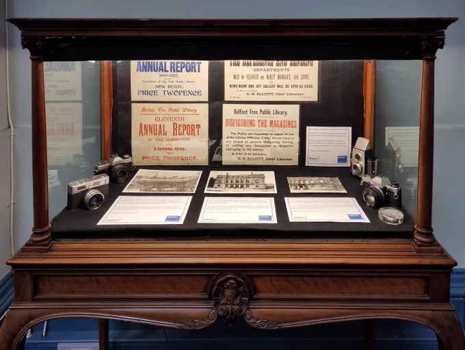 Wooden display case showcasing vintage cameras, historical Belfast Free Public Library annual reports, and old photographs of library buildings.