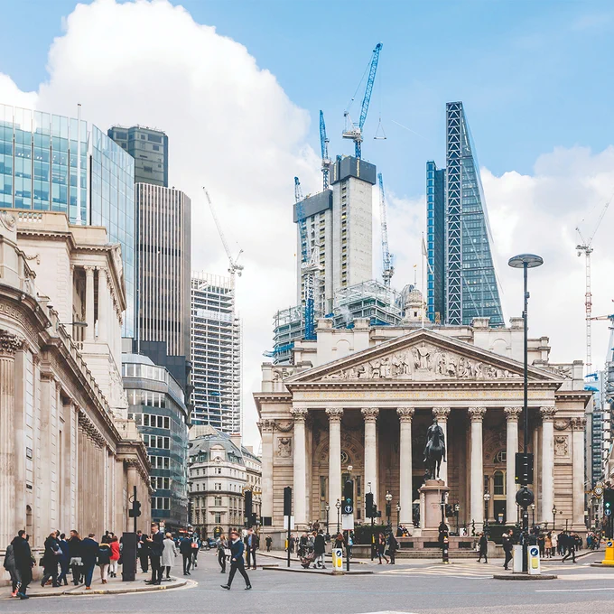 The London stock exhange building with the Bank of England to the right and modern sky scrapers in the background. The street in front is busy with people walking.