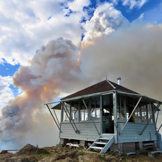 Big smoke cloud billows above a fire lookout structure, with blue sky and regular clouds over head.