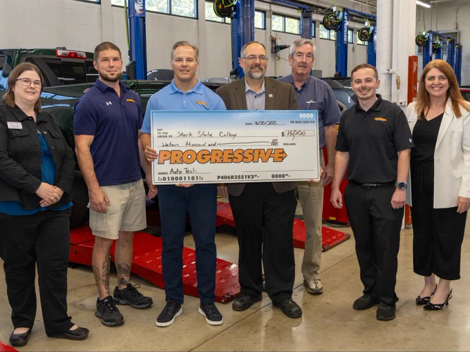Group of seven people posing with a large check from Progressive in an automotive technology school.