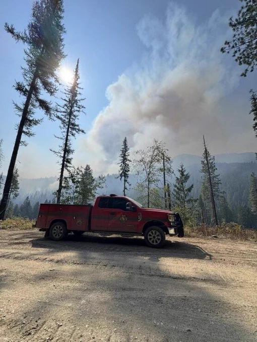 Parked firetruck in foreground with large smoke cloud from forest fire in the distant background.