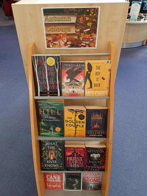 Photograph of a tall shelf of books with tiered shelves. A poster at the top of the shelf reads "Autumn Colours". Book titles include The Last House On Needless Street by Catriona Ward, The Boy Is Back by Meg Cabot, A Change of Circumstance by Susan Hill, and The House at Phantom Park by Graham Masterton.
