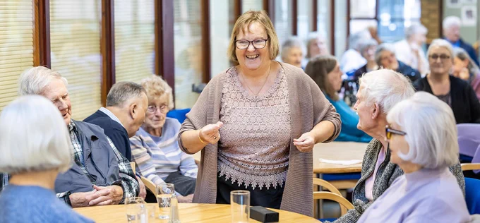 A group of older adults seated around tables at a Runnymede social centre. A person stands in the middle, engaging with the group. Tables have water glasses and personal items. The room has large windows and a bright, welcoming atmosphere.