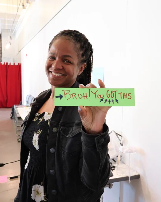 Smiling woman holds a bright green sign reading "Bruh! You GOT THIS" with arrows pointing to the text, conveying encouragement.