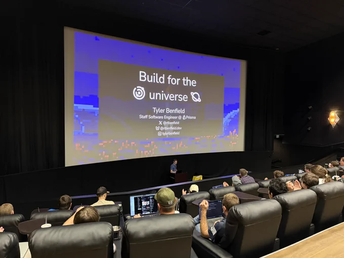 A speaker presents in a movie theater-style room with reclining seats filled with attendees. On the large projector screen is a slide that reads: “Build for the universe” with contact info for Tyler Benfield, Staff Software Engineer at Prisma. His social handles are shown: @rtbenfield on X, rtbenfield.dev, and tylerbenfield on GitHub.
