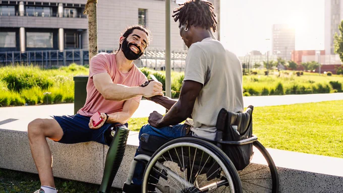Two men shaking hands in an outdoor setting. One man is using a prosthesis and the other is using a wheelchair. They are smiling and engaging in conversation.