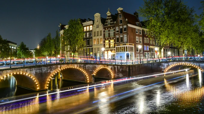 A scene of one of Amsterdam's canal bridges by night. There is motion blur on the image from the lights on the canal boats passing by.