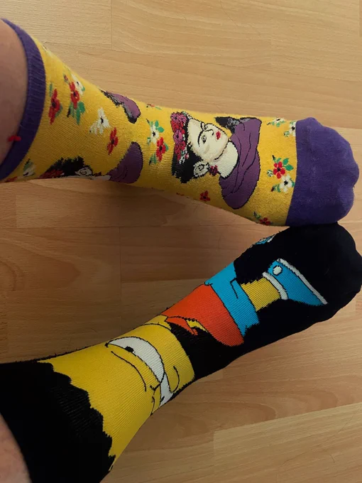 A photo of two feet on which a pair odd socks being worn - one with a Bart Simpson design and one of Frida Kahlo.
