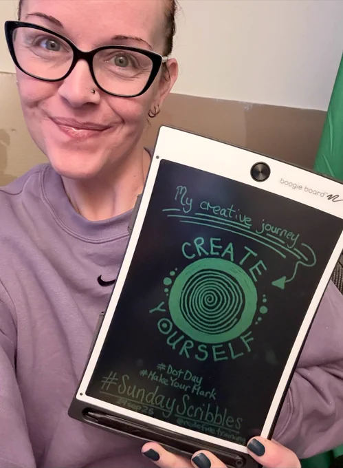 A smiling selfie of Georgina Dean, wearing a purple sweatshirt and cat-eye glasses, holding up a Boogie Board. The board displays her hand-drawn scribble for #SundayScribbles, which features a large, spiraling dot with the words "CREATE YOURSELF" encircled around it to celebrate #DotDay.