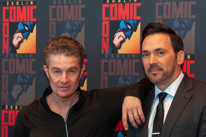 The image shows two men posing in front of a backdrop adorned with a repeated graphic design. One man, on the left (James), has curly brown hair and is wearing a black zip-up jacket. He is slightly leaning with his arm resting on the shoulder of the second man (Jason). The man on the right has short dark hair, a trimmed beard, and is wearing a striped suit with a white shirt and dark tie. The backdrop they are standing against is decorated with the bold red text "DUBLIN COMIC CON" and features artwork of a stylized superhero's face in a blue and orange color scheme.