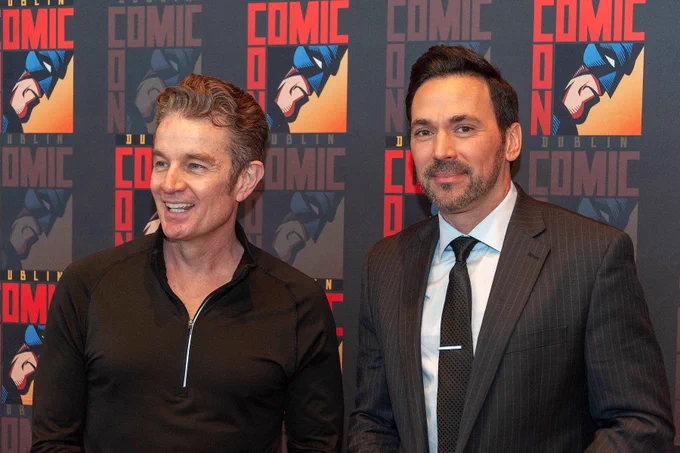 The image features two men standing side by side in front of a backdrop with a repeated pattern. The backdrop prominently displays the words "COMIC CON" in bold red letters and an illustration of a masked superhero head in blue and red tones. The word "DUBLIN" appears beneath the "COMIC CON" text. The man on the left is wearing a black zip-up shirt, while the man on the right is dressed in a pinstripe suit with a white shirt and a dark tie. They both appear to be smiling, creating a friendly and welcoming atmosphere.
