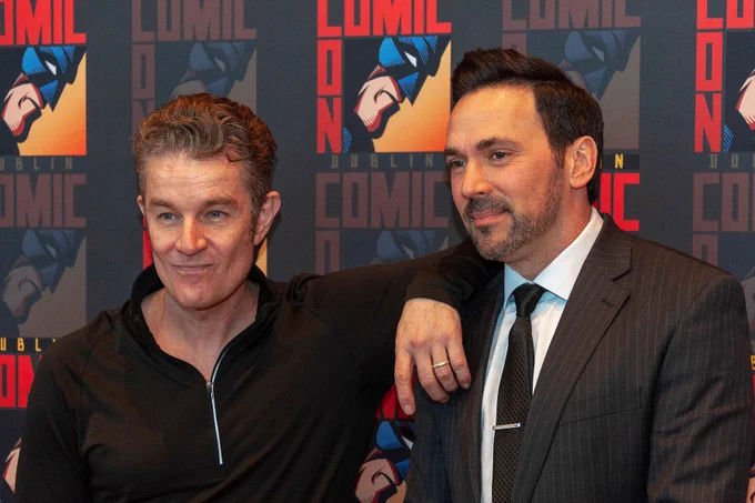 The image features two men posing in front of a backdrop promoting a Comic Con event in Dublin. The man on the left has short, curly hair and is wearing a black zip-up top. He is smiling and has his arm around the shoulders of the man on the right. The man on the right has short, dark, slicked-back hair and a trimmed beard, wearing a pinstripe suit with a white shirt and black tie. The backdrop is decorated with repeated comic-style graphics of a masked character and the words "Comic Con Dublin" in bold red and yellow text.
