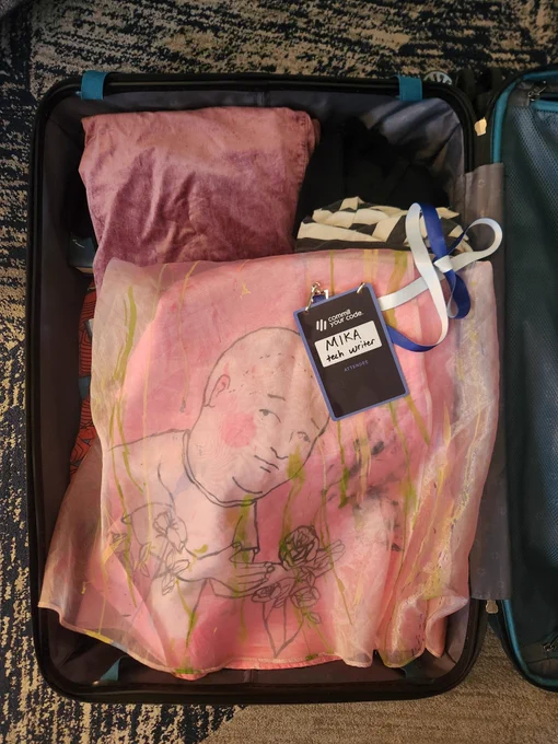 An open suitcase with folded clothes inside, including a hand-painted skirt with the likeness of Bobby Hill from King of the Hill. A tech conference name tag lies on top, and it says MIKA - tech writer