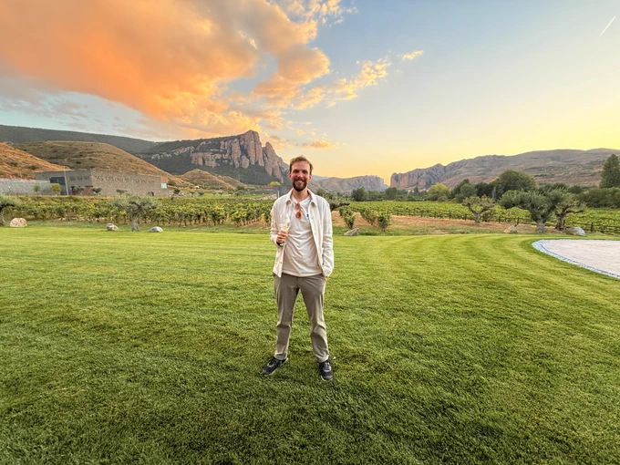 Me standing center on green grass with grapevines and mountains in the background. Its sunset, s the sky is turning from blue to yellow and the clouds are turning orange.