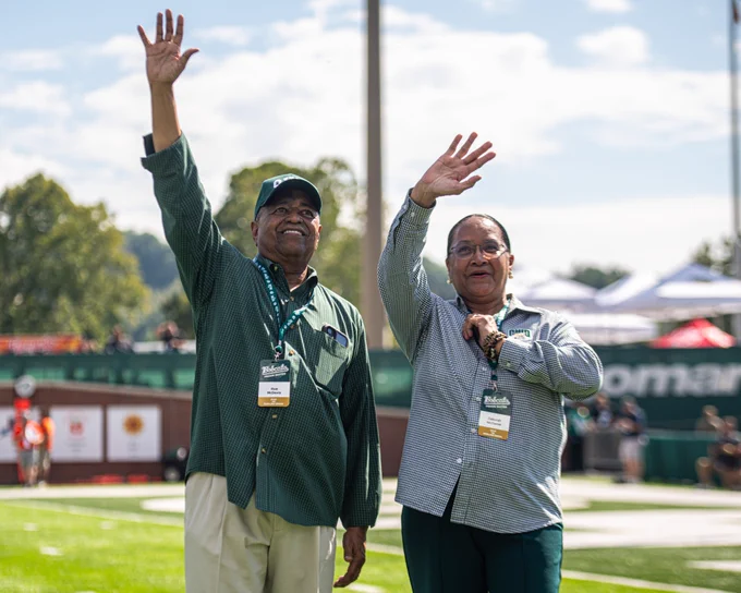 Two people wave and smile while on football field.