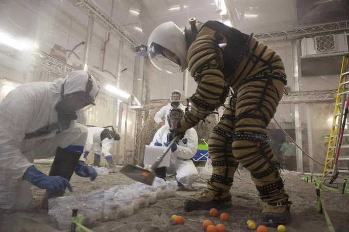 Two individuals in protective suits examining materials in a laboratory setting, with one wearing an advanced, bulky suit and handling equipment, amidst a backdrop of construction or renovation work.