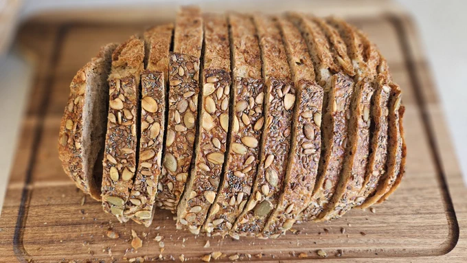 A photo of a seeded loaf, sliced by hand on a wooden bread board.