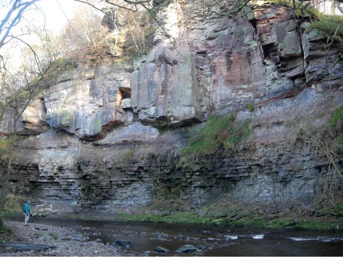 A person stands next to a river. Behind the river is a rocky cliff face showing different sedimentary layers of various thicknesses and textures.
