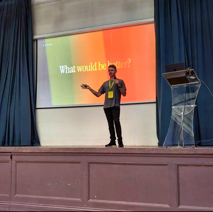 John O'Nolan on stage at LoopConf in front of a slide that says "What would be better?"