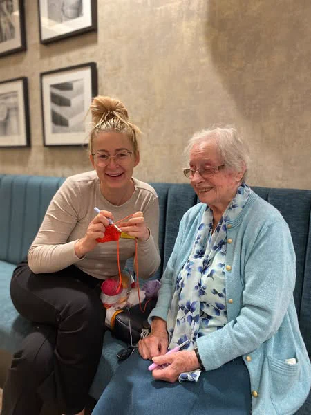 Two people sit on a blue bench. One sits crocheting with yarn and needles. The other wears a light blue cardigan and patterned scarf. Framed pictures hang on the wall behind them.