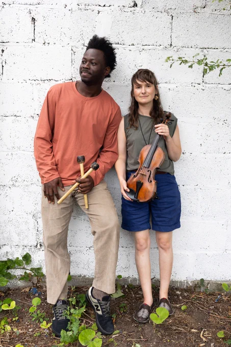 A man in a peach coloured t-shirt and a woman in a green vest are leaning against an exterior wall holding musical instruments. He is holding balafon mallets and she is holding a violin.