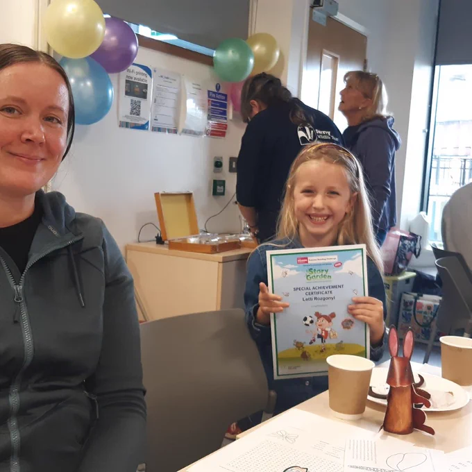 Photo of a little blond girl who is smiling, holding here Summer Reading Challenge certificate - her mother is sitting to her right and is also smiling. There are other people in the background and balloons on the wall.