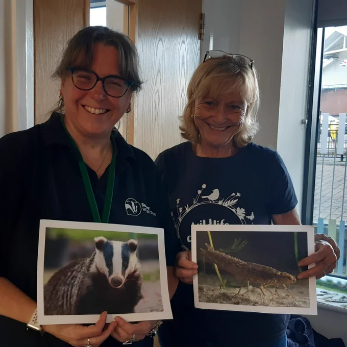 Photo of two smiling women - the one on the left is holding a photo of a badger and the woman on the right is holding a photo of a shrimp underwater