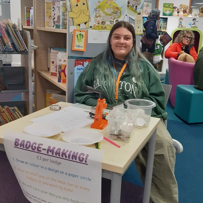 Photo of a smiling woman in a green sweatshirt, sitting behind a table in a library - the poster on the front of the table reads "badge making". You can see people in the background