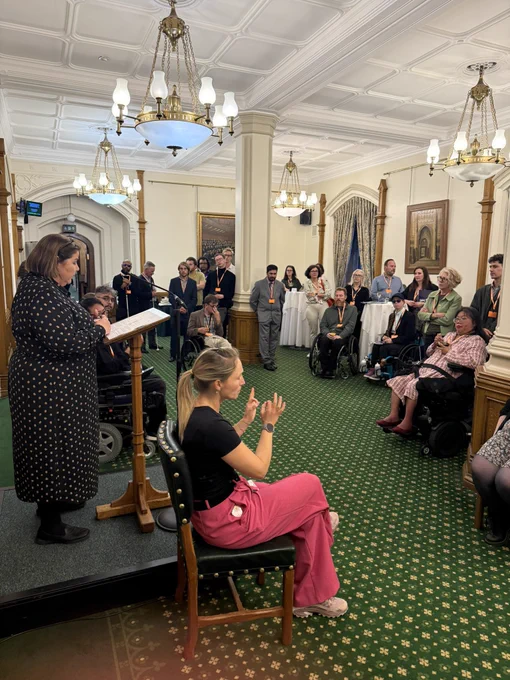 Julie Minns MP speaks at a podium in the Churchill Room, Houses of Parliament, while James Lee, the event chair, sits in a wheelchair beside her. Other panel members are seated nearby