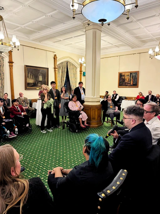 Audience and panel in the Churchill Room, Houses of Parliament. A female guest in a wheelchair and pink dress asks a question while panelists, including Gáibhin, Ria, Michelle, and Mike, listen and respond under chandeliers and paintings.