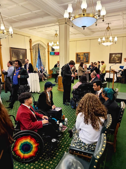 Busy scene in the Churchill Room, Houses of Parliament, with groups of people networking and talking. Several wheelchair users and seated guests are present, surrounded by chandeliers, paintings, and green patterned carpet