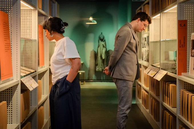 Two people viewing an exhibition designed to look like an archive, with shelving and boxes.
