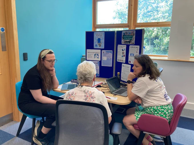 Three people sat round a table talking, with a laptop in front of them. On the table there is a notice board with posters about digital skills.
