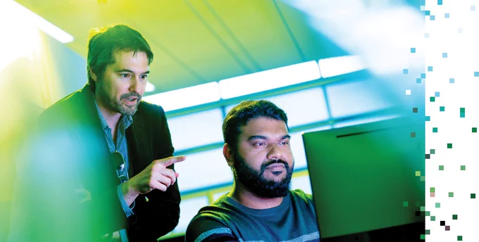 A professor stands behind a student who is sitting at a computer 