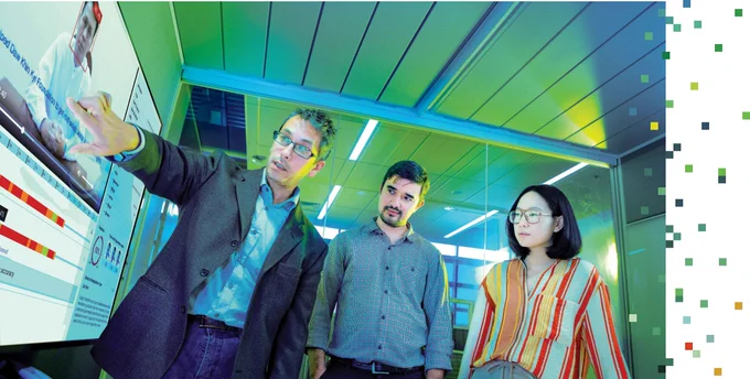 A professor points at a screen while standing next to two students 