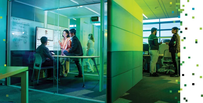 Professor and two students analyzing data on a screen inside a small glass walled cubicle meeting space. Other people stand outside the cube in a larger lab room chatting