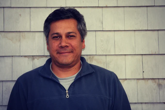 Man wearing a navy blue zip-up jacket standing against a weathered wooden shingle wall outdoors.
