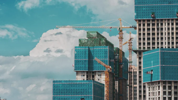 Image of a construction site with multiple cranes and several high-rise buildings under construction against a backdrop of a clear blue sky with scattered clouds.