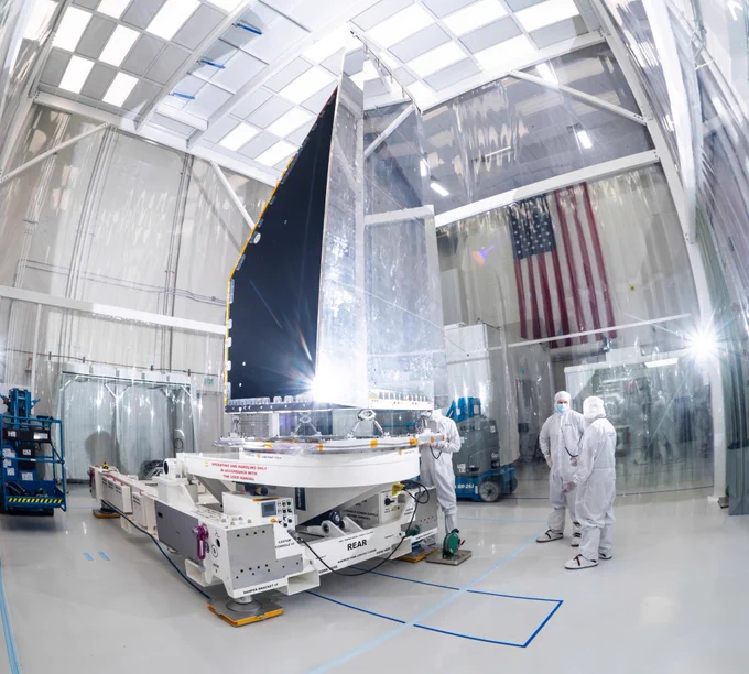 The NEO Surveyor instrument enclosure is shown on its side in a bright, white clean room. The mirrored side reflects the room and the light from the camera while the dark sides are shown. Two people in bunny suits stand near the spacecraft and in the background outside of the room is an American flag draped against a wall.

Credit: Space Dynamics Laboratory/Allison Bills