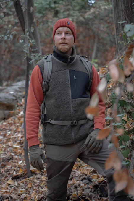 A man stands in a forested area with autumn leaves on the ground, wearing a red beanie, red sweatshirt, brown fleece vest, and outdoor trousers. He has a backpack and gloves, looking slightly to the side with a calm expression.
