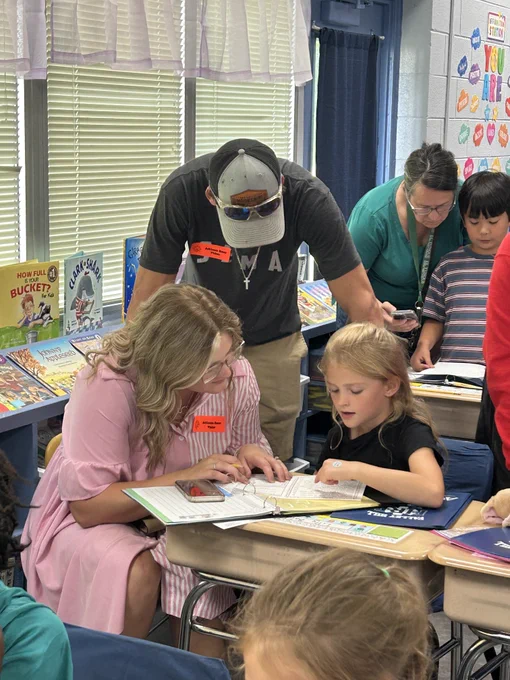 second grade girl sits at her seat. her mom sits next to her and leans over her desk. dad stands behind them and hunches over to participate