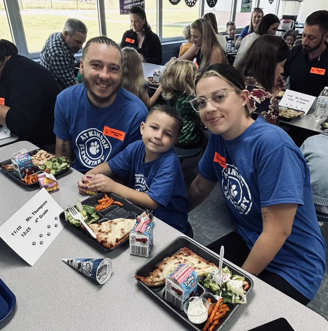 kindergarten boy sits in between his mom and dad at lunch. they are all smiling and are sitting at the cafeteria tables. there are more families behind them.